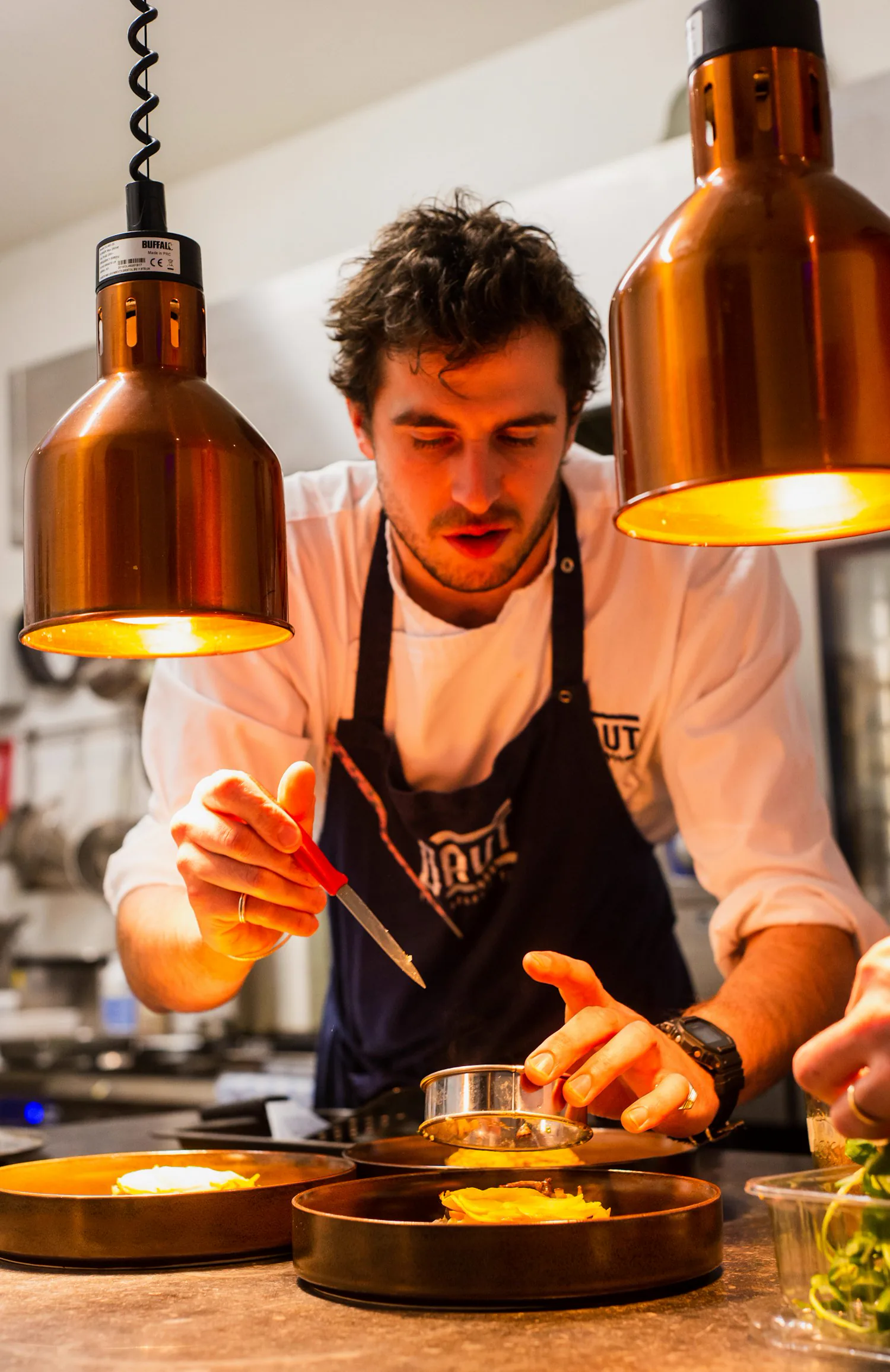 A chef in a dark apron holding a knife, working under warm copper pendant lights